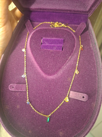 A delicate gold chain necklace with small colorful charms, displayed inside an open purple velvet jewelry box.